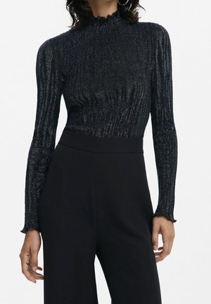 Femme portant un haut noir ajusté à manches longues scintillant avec un col et des poignets à volants, associé à un pantalon noir taille haute.