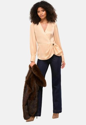 Beige Wickelbluse mit langen Ärmeln, tailliertem Schnitt und seitlichem Schleifendetail. Kombiniert mit dunkelblauen weit geschnittenen Jeans und einem braunen Pelzschal.