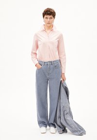 Lichtroze overhemd met puntkraag, gecombineerd met losse blauwe spijkerbroek en een grijze spijkerjas over één arm gedrapeerd.