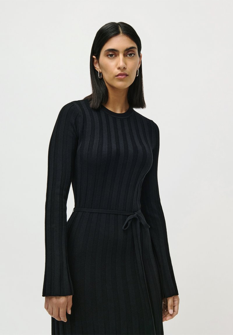Donna con capelli neri alla spalla che indossa un vestito aderente nero a costine con una cintura annodata in vita, in piedi su uno sfondo semplice.