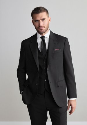 Mies, jolla on lyhyet hiukset ja parta, pukeutuneena mustaan kolmi-osaisen puvun, mustan solmion, valkoisen paidan ja burgundin taskuliinan kanssa, seisomassa käsi taskussa.