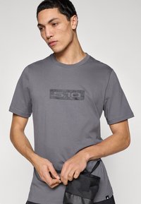 Γκρι βαμβακερό t-shirt με κοντά μανίκια, με υφασμένο μαύρο ορθογώνιο λογότυπο στο στήθος. Το μοντέλο κρατάει μια μαύρη θήκη.