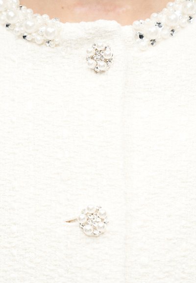 Λευκό υφασμάτινο ύφασμα με κουμπιά από πέρλες και στρας και διακοσμητικό γιακά με πέρλες σε ρούχο.