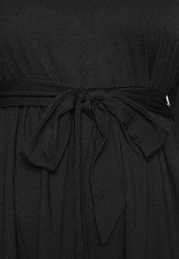 Vestido preto com um tecido de malha texturada, corpete justo e uma faixa de laço na cintura, criando um efeito drapeado.