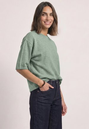Femme aux cheveux bruns mi-longs portant un haut vert à manches courtes et un jean bleu foncé, souriante avec une main dans la poche du jean.