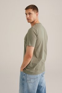 Camiseta de algodón verde oliva de cuello redondo con mangas cortas, corte relajado y costuras detalladas. Combinada con pantalones cortos de mezclilla azul claro.