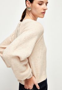 Helle beige gestrickte Pullover mit Ballonärmeln und einem Rundhalsausschnitt. Weiche Textur und enganliegender Saum. Das Model trägt einen dekorativen Ohrring.