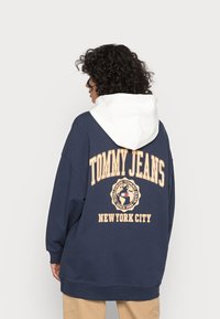 Námornícky modrá oversized mikina s bielou kapucňou. Obsahuje nápis "TOMMY JEANS" a kruhové logo v béžovej farbe. Mäkko tkaná látka a voľný strih.