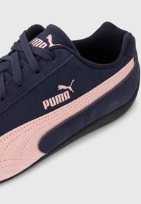 Σκούρο μπλε αθλητικό παπούτσι Puma με ροζ ρίγα και λογότυπο, υφή από καστόρι, σχεδιασμός με κορδόνια και μαύρη σόλα, τοποθετημένο σε γωνία για να φαίνονται τα πλευρικά και επάνω μέρη.