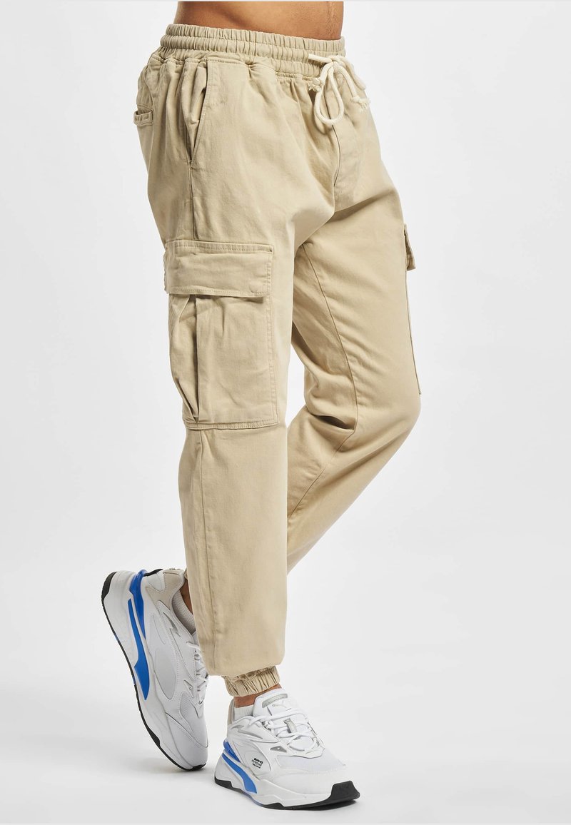 Beige cargojoggingbroek met elastische boorden en een taille met trekkoord, gedragen met witte en blauwe sportieve sneakers.