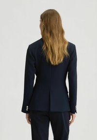 Navy tailored blazer with structured fit, featuring a single vent, long sleeves, and silver buttons on cuffs. Smooth fabric texture.