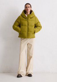 Olivgrüne Pufferjacke mit Kapuze, ausgestattet mit einem Reißverschluss vorne, weiten Ärmeln und horizontalem Steppdesign. Kombiniert mit beigen Cordhosen.