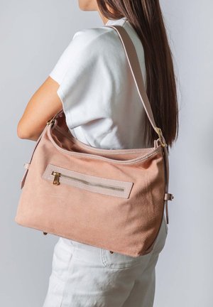 Vrouw met een lichtbruine suède schoudertas met een voorvak met rits, gekleed in een wit shirt en een witte broek, zijwaarts kijkend.