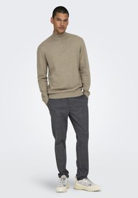 Beige Strick-Polo-Sweater mit Kragen und Reißverschluss, kombiniert mit grauen Hosen im Karomuster und hellen Sneakern. Schlichtes und modernes Design.
