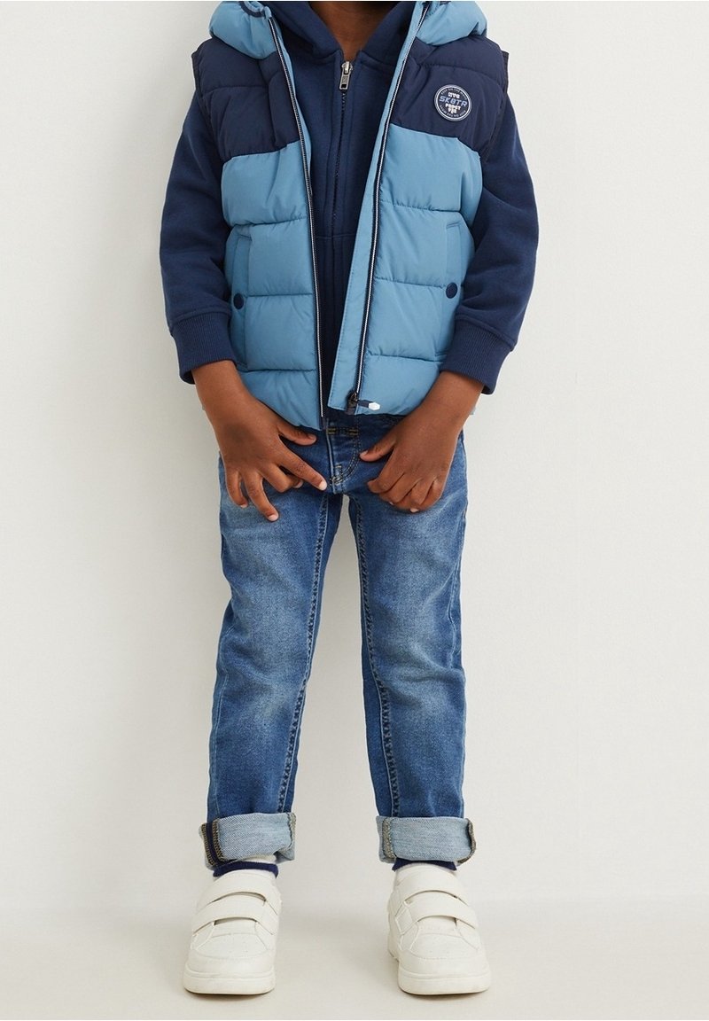 Enfant portant une doudoune bleue par-dessus un sweat à capuche bleu marine, un jean bleu retroussé et des baskets blanches, debout devant un fond uni.