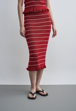 Rød, horisontalt stripet midiskjørt med rysjekant, brukt med svarte flip-flops, vist fra midt på overkroppen til føttene på grått gulv.