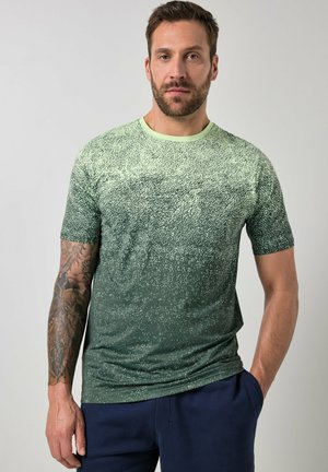 Mężczyzna z brodą, ubrany w zieloną koszulkę z gradientowym wzorem i granatowe spodnie, stoi z jedną ręką w kieszeni na jednolitym tle.