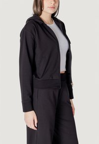 Felpa nera con zip, dal tessuto morbido, tasche frontali e polsini a costine, abbinata a una maglietta bianca aderente. Presenta un design classico con cappuccio.