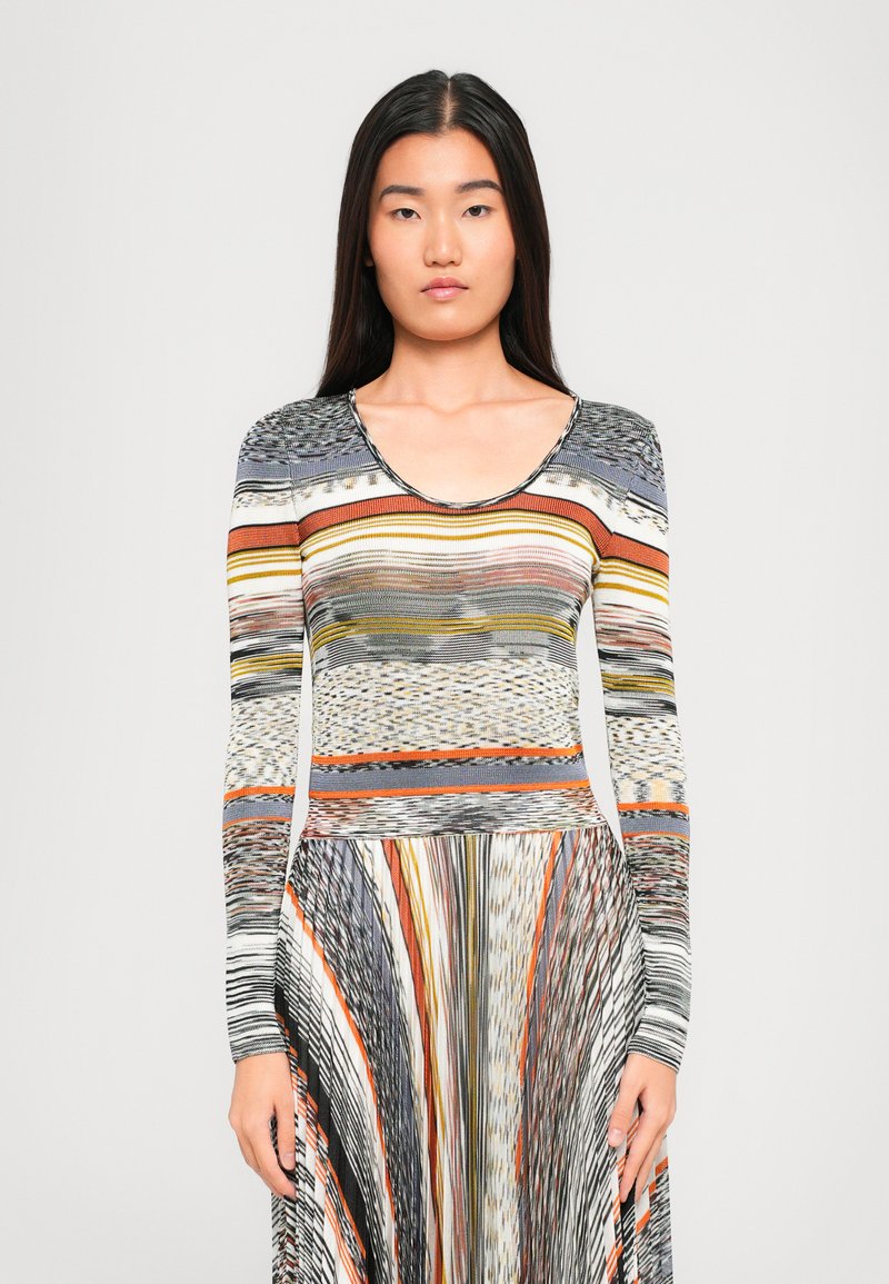 Giovane donna con lunghi capelli scuri che indossa un vestito a maniche lunghe a strisce multicolori con vari motivi, rivolta verso l'avanti su uno sfondo bianco.