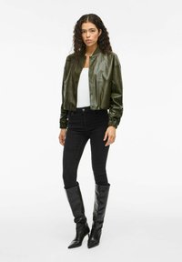 Grüne Lederbomberjacke mit taillierter Passform, elastischen Bündchen und vorderen Druckknöpfen, kombiniert mit schwarzen engen Jeans und hohen schwarzen Stiefeln.