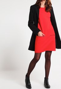 Rode jurk met korte mouwen en een rechte snit, met ritszakken. Gecombineerd met een zwarte jas en zwarte enkellaarsjes. Glad textuur.