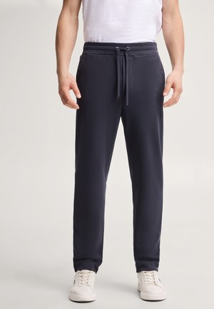 Person trägt dunkelblaue Jogginghose mit Kordelzug in der Taille und weiße Sneakers, steht mit locker an den Seiten hängenden Händen.