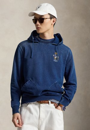 Fiatal férfi kék kapucnis pulóverben, melyen egy plüssmackó grafika látható, fehér sapkában, sötét napszemüvegben, fehér nadrágban és fonott bőr övben.