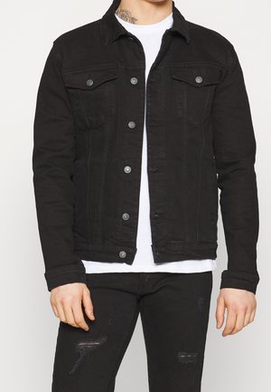 Mann trägt eine schwarze, geknöpfte Jeansjacke über einem weißen T-Shirt mit zerrissenen schwarzen Jeans und zeigt ein Hals-Tattoo mit floralem Design.