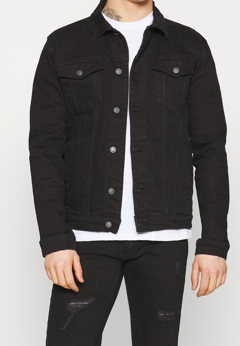 Homme portant une veste en jean noire boutonnée sur un t-shirt blanc avec un jean noir déchiré, montrant un tatouage au cou avec un motif floral.