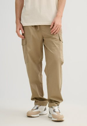 Hombre con pantalones cargo beige y zapatillas blancas, de pie con las manos relajadas a los lados contra un fondo liso.