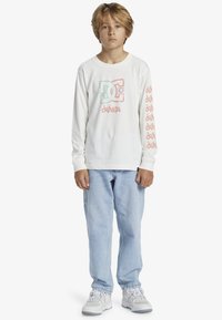 Weißes Langarm-T-Shirt mit buntem Grafiklogo auf der Brust und wiederholtem Text entlang der Ärmel, kombiniert mit hellblauen Jeans und Sneakers.