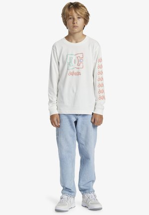 Weißes Langarm-T-Shirt mit buntem Grafiklogo auf der Brust und wiederholtem Text entlang der Ärmel, kombiniert mit hellblauen Jeans und Sneakers.