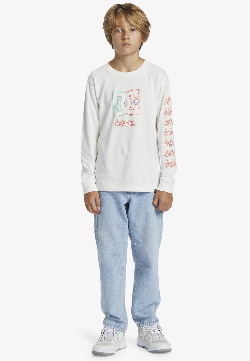 Weißes Langarm-T-Shirt mit buntem Grafiklogo auf der Brust und wiederholtem Text entlang der Ärmel, kombiniert mit hellblauen Jeans und Sneakers.