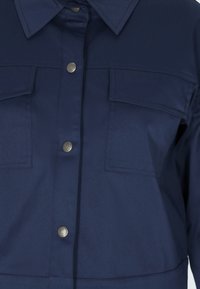 Námořnická modrá košile s knoflíky, límečkem, dvěma náprsními kapsami a stříbrnými patentkami. Hladká textura a přizpůsobený design.