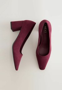 Zapatos de tacón alto en ante sintético burdeos con puntera afilada y tacón grueso. La suela presenta un ribete negro. Forro interior plano y suave.