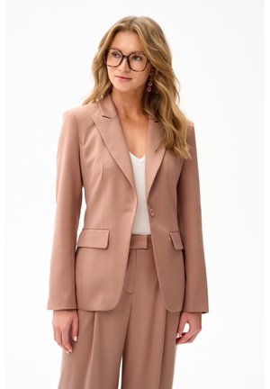 Vrouw met golvend blond haar, die oversized schildpadbril draagt, een rozenkleurig pak en een wit topje, staand tegen een witte achtergrond.