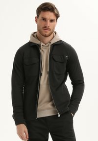 Uomo con felpa beige indossata sotto un giubbotto nero con zip e tasche a pattina, in piedi con una mano in tasca contro uno sfondo bianco.
