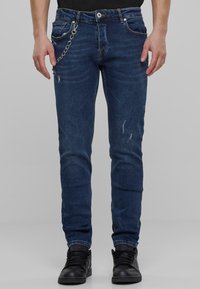 Kék farmernadrág, slim fit szabással, megkopott részletekkel és ezüst lánc díszítéssel. A kezek az oldalon, fekete sportcipőt viselve.