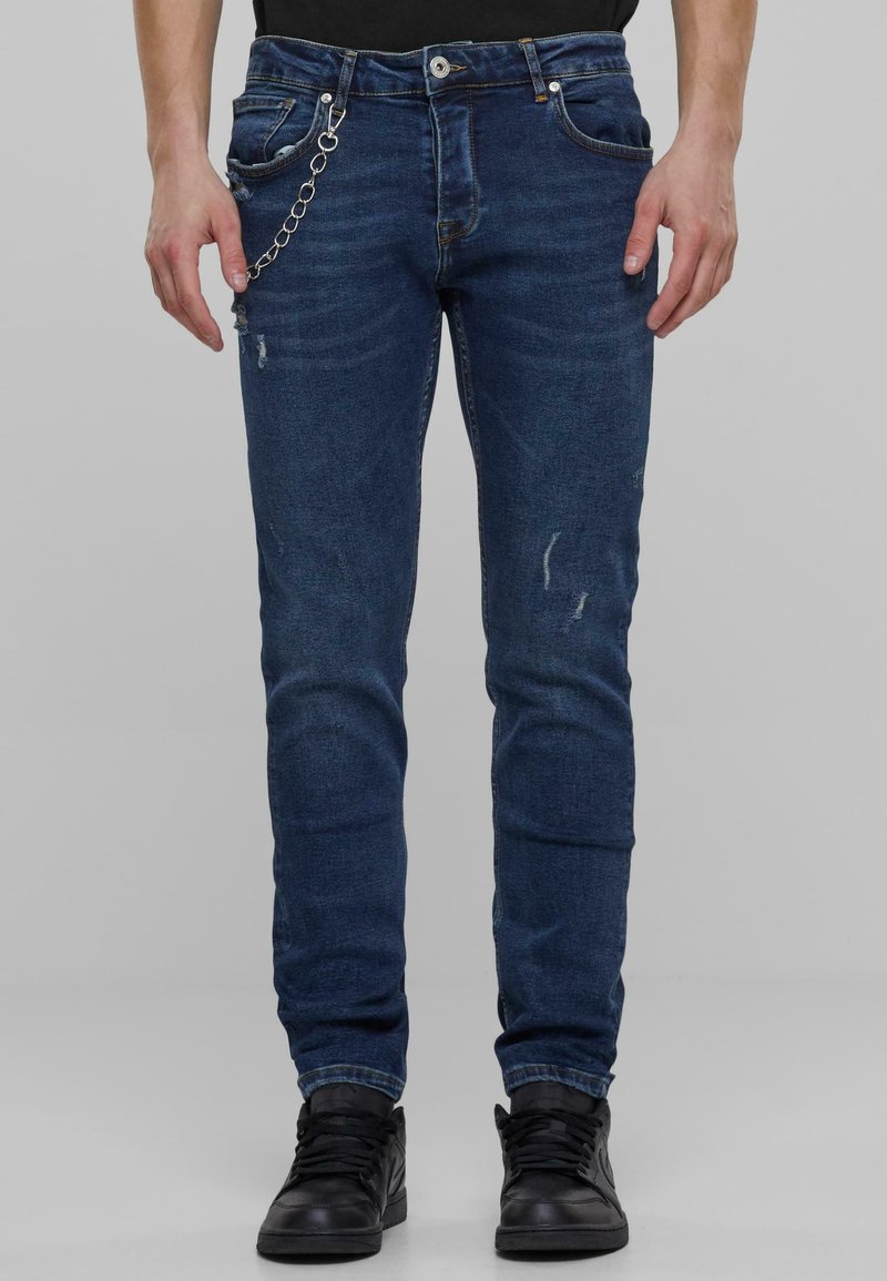 Kék farmernadrág, slim fit szabással, megkopott részletekkel és ezüst lánc díszítéssel. A kezek az oldalon, fekete sportcipőt viselve.