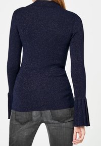 Marineblauer Strickpullover mit körperbetontem Schnitt und ausgestellt geschnittenen Ärmeln, der subtilen metallischen Glitzer für Textur aufweist. Kombiniert mit dunkelgrauen Jeans.