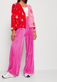 Cardigan rouge et rose avec des motifs colorés, assorti à un pantalon large plissé rose et des baskets blanches. Couleurs vives et textures distinctes.