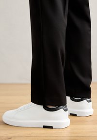 Fehér sneaker fekete sarokbetétekkel, sima bőr felsőrésze, lekerekített orra és lapos talpa van, fekete, szűkített nadrággal párosítva, fából készült padlón.