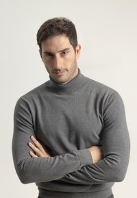 Hombre con cabello corto y oscuro y barba de unos días, vistiendo un suéter de cuello alto gris, mirando directamente con los brazos cruzados frente a un fondo sencillo.