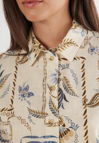 Chemise de couleur claire avec un motif floral et de feuilles en bleu et marron. Présente un col pointu et une patte de boutonnage avec des boutons ronds.