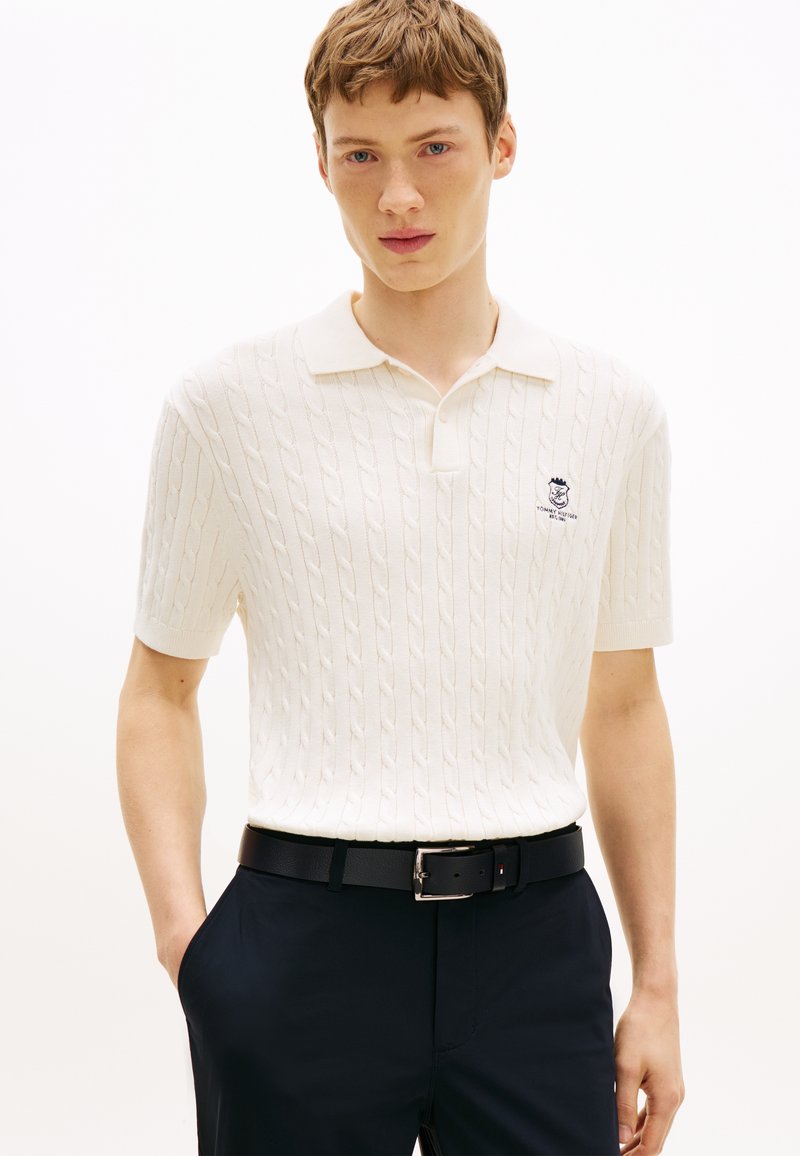 Νέος άνδρας που φοράει λευκό πλεκτό πόλο με κοντά μανίκια και κεντημένο λογότυπο, μαύρη ζώνη και σκούρο παντελόνι, στέκεται μπροστά σε λευκό φόντο.