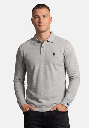 Junger Mann trägt ein hellgraues Polohemd mit langen Ärmeln und einem kleinen dunklen Logo auf der Brust, Hände in den Taschen, vor einem schlichten Hintergrund.