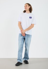 Hvit bomulls T-skjorte med et kvadratisk blått logo på brystet, sammen med lyseblå rettskårne jeans og svarte joggesko.