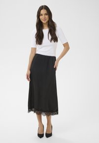 Jupe longue noire avec bordure en dentelle à l'ourlet, associée à un haut blanc ajusté à manches courtes. Le mannequin porte des talons hauts noirs.