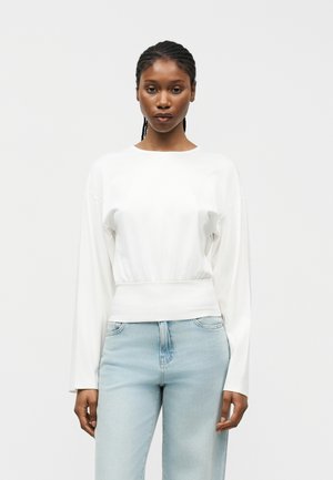 Γυναίκα με πλεγμένα μαλλιά φοράει ένα μακρυμάνικο λευκό τοπ και ανοιχτό μπλε τζιν, όρθια μπροστά σε ένα απλό ανοιχτό γκρι φόντο.