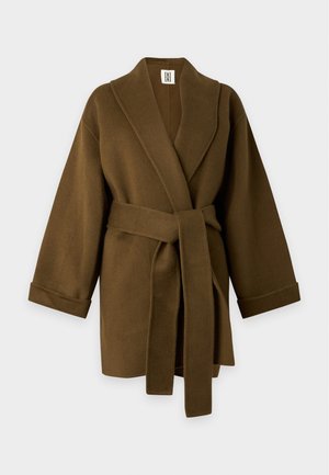 Kvinde iført en brun oversized frakke, hvid top, mørkebrun midinederdel med frontslids og sorte læderstøvler, der står på en ensfarvet baggrund.